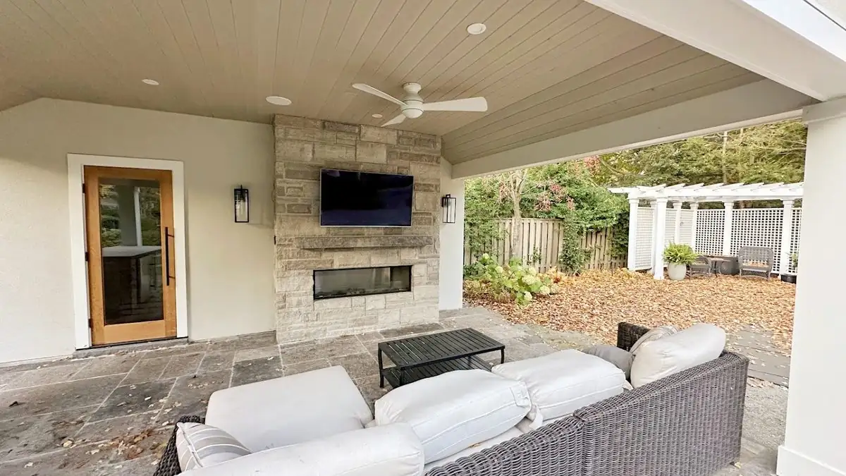 Covered outdoor living room with a TV above a stone fireplace, in-ceiling speakers and a ceiling fan over a sectional