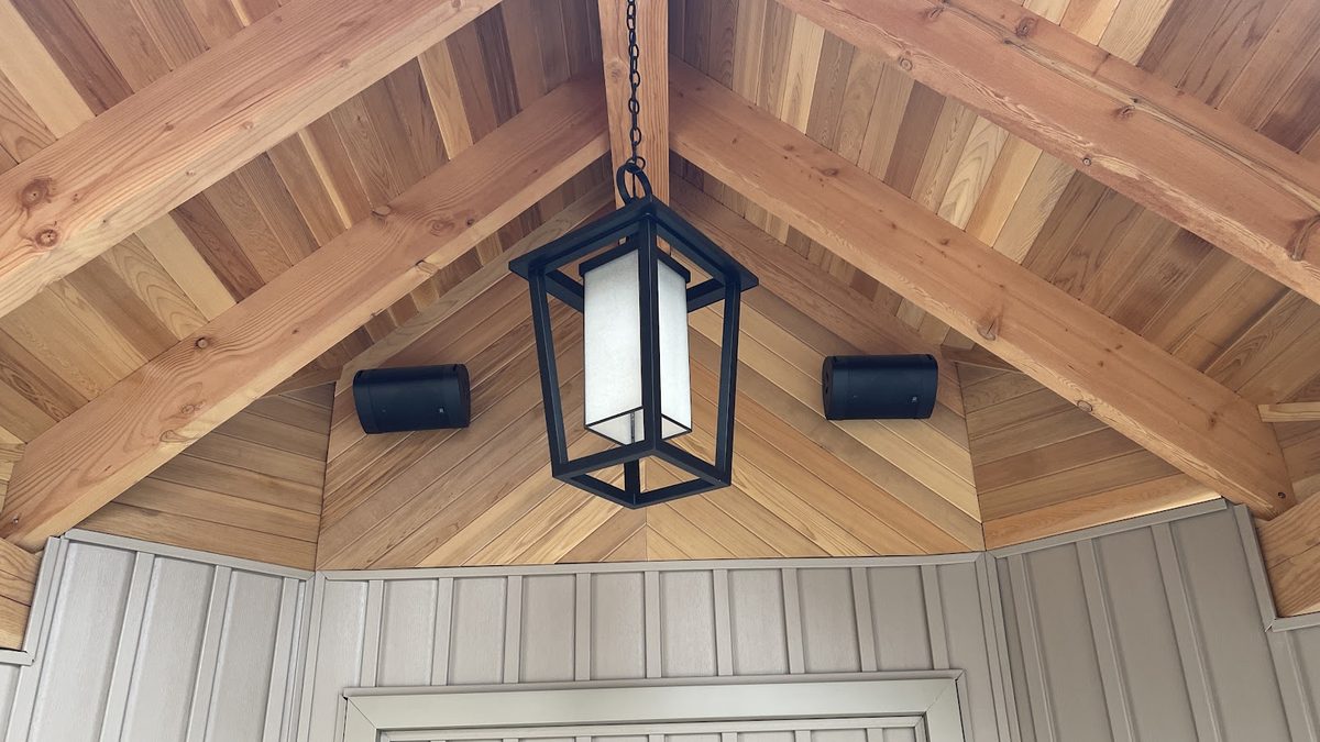 Two outdoor speakers mounted under a cedar porch ceiling beside a hanging lantern